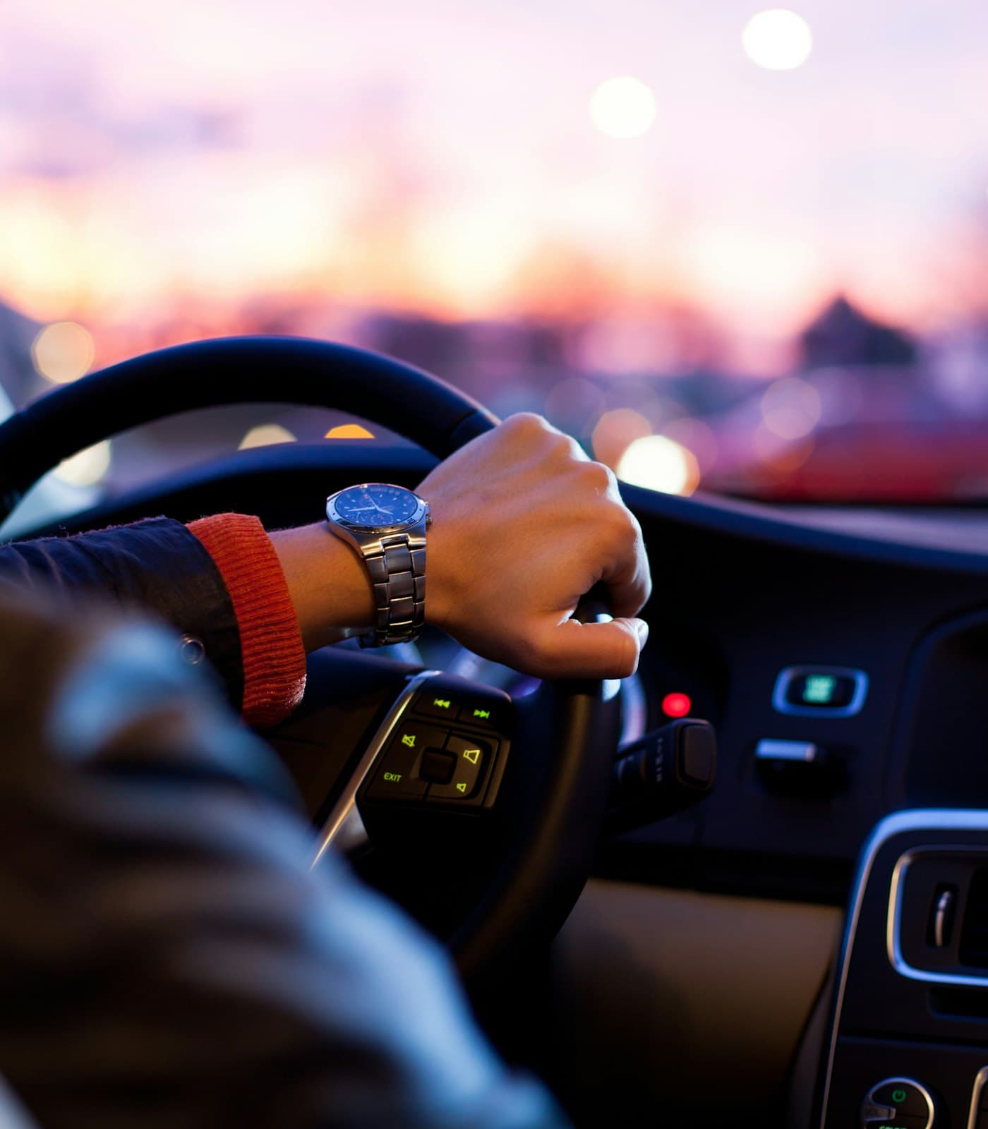 Driver's hand on a steering wheel with city lights blurred through the windscreen